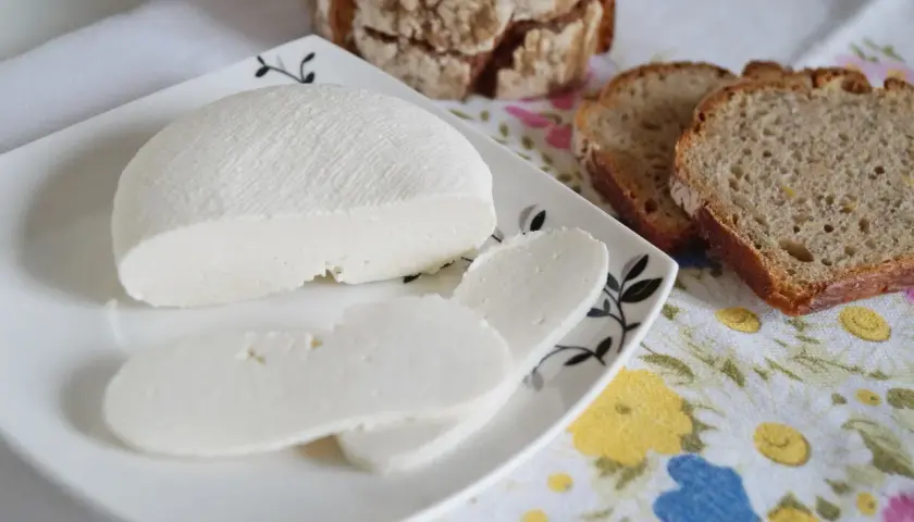 Świeży ser typu bufala, pokrojony w plastry, obok kromek chleba na kwiecistym obrusie.