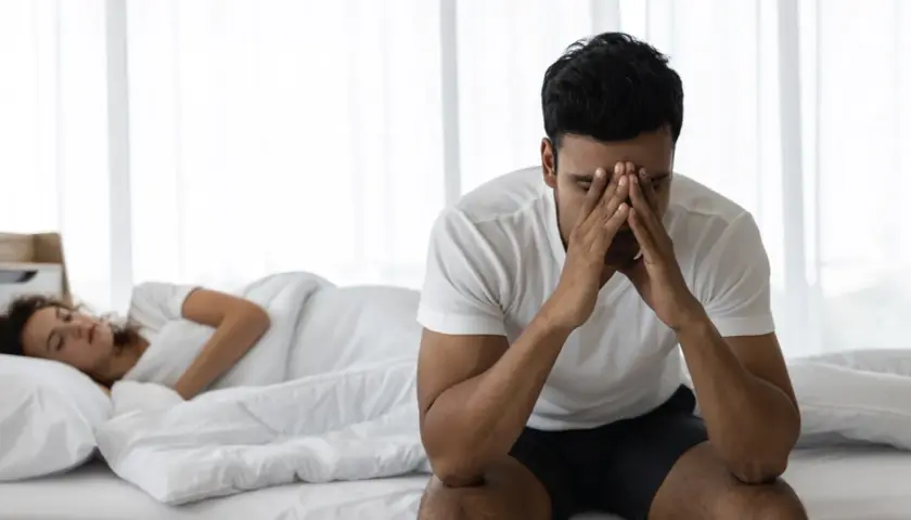 Uomo seduto sul letto con le mani sul viso, preoccupato per l'eiaculazione precoce. La donna dorme nel letto alle sue spalle.