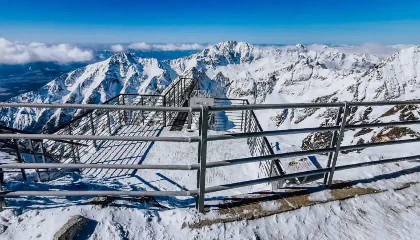 Widok z tarasu widokowego na szczycie Łomnicy. Stalowe balustrady i schody prowadzą do zapierającej dech w piersiach panoramy ośnieżonych gór.