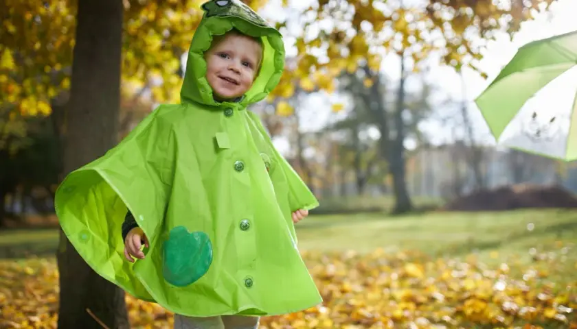 Uśmiechnięty maluch w zielonej pelerynie z kapturem w kształcie żaby, idealnej na jesienne zabawy. Dowiedz się, jak uszyć pelerynę dla dziecka!