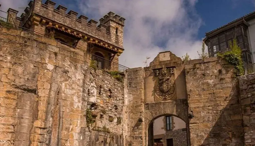 Antigua muralla y arco de entrada en Fuenterrabía, con un escudo nobiliario y un cielo azul con nubes.