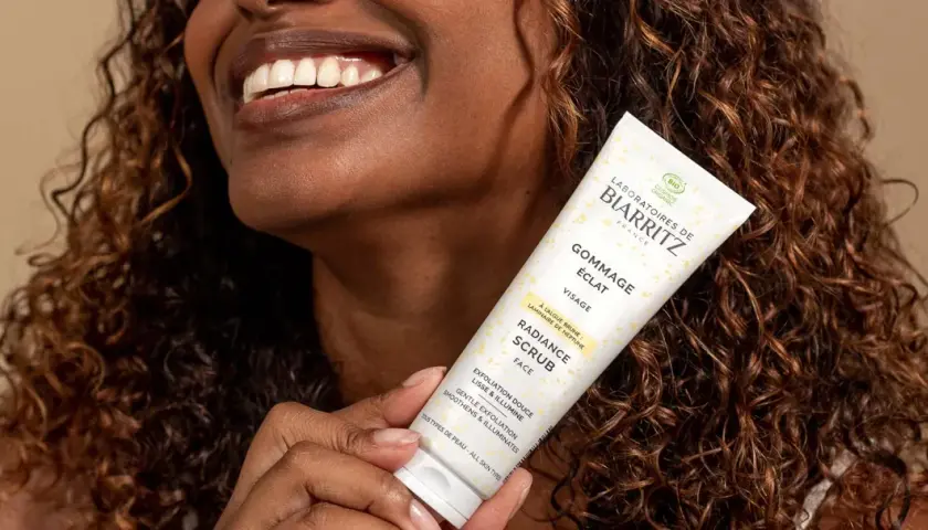 Une femme souriante tient un tube de gommage visage éclat. Ce produit sert à lisser et illuminer la peau, révélant sa beauté naturelle.
