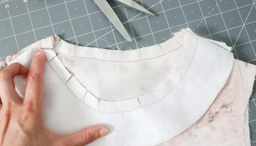 A hand guides the inner facing of a garment, showing the clipped seam allowance for a smooth neckline finish.