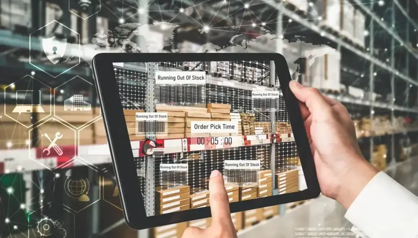 Ręce trzymają tablet z widokiem magazynu, pokazujący alerty "Running Out Of Stock" i czas kompletacji zamówienia. To przykład nowoczesnego zarządzania łańcuchem dostaw.