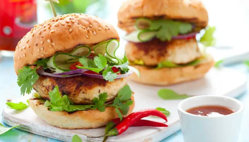 Dos hamburguesas de pollo frescas y jugosas, adornadas con pepino, cebolla morada, cilantro y chile rojo.