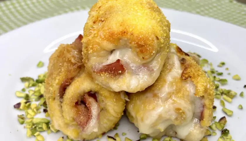 Tre involtini di pollo dorati e ripieni di formaggio filante e prosciutto, adagiati su un letto di pistacchi tritati.