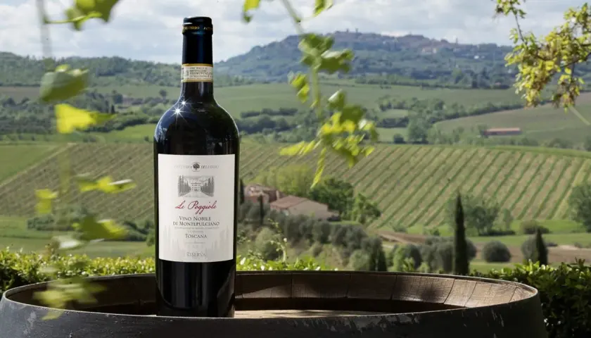 Bottiglia di Vino Nobile di Montepulciano su una botte, con vigneti e colline toscane sullo sfondo. Un invito a una visita cantina Montepulciano.