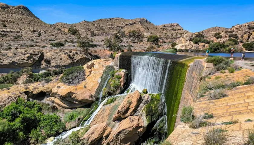 Cascada en el pantano de Elche, con rocas cubiertas de musgo y vegetación verde. Personas observan el agua.