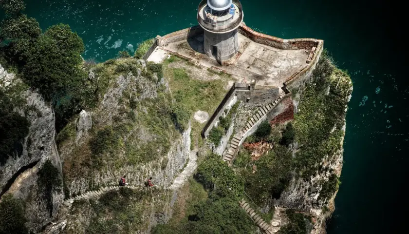 Dos personas caminan por un sendero empinado hacia el faro de caballo, un faro solitario en un acantilado rocoso sobre el mar turquesa.