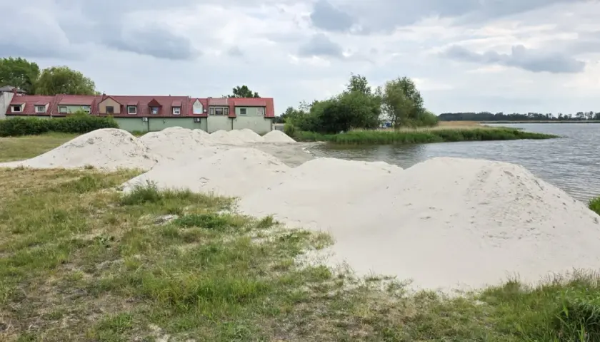 Piasek na plaży w Kamieniu Pomorskim czeka na budowę. W tle zielone budynki i spokojna woda.