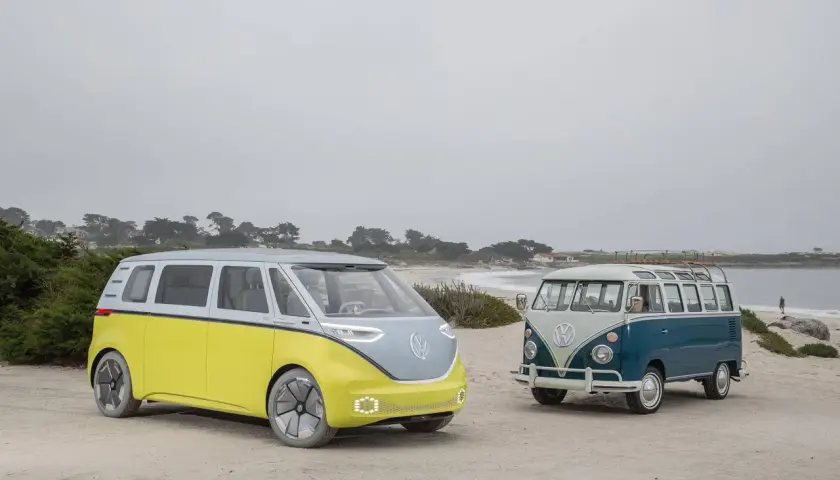 Deux combi VW, un moderne jaune et gris, l'autre vintage bleu et blanc, sur une plage brumeuse.