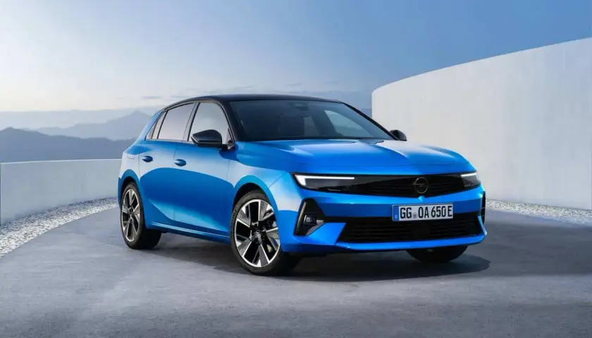 Błękitny opel elektryczny z czarnym dachem, zaparkowany na betonowej nawierzchni, z górzystym krajobrazem w tle.