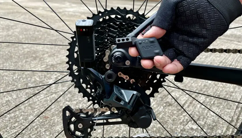 Close-up of a mountain bike's rear derailleur, showing the NX5 electronic shifting system being adjusted by a gloved hand.