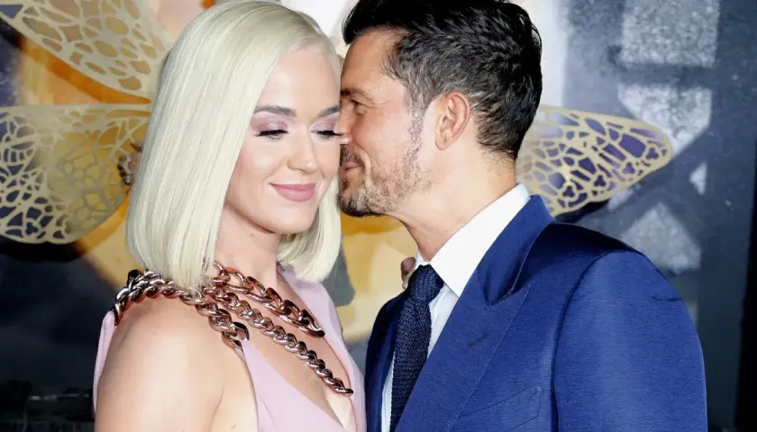 Orlando Bloom szepcze coś do ucha Katy Perry, która ma na sobie różową sukienkę i gruby, złoty naszyjnik.