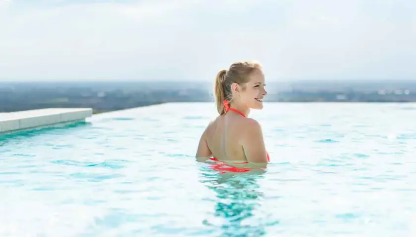 Frau genießt das Schwimmen in einem Infinity Pool, der über den Horizont zu schweben scheint. Was ist ein Infinity Pool? Ein Pool, dessen Wasser über den Rand zu fließen scheint und eine Illusion von grenzenlosem Wasser erzeugt.