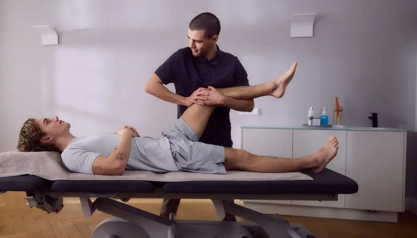 Physiotherapeut dehnt das Bein eines Patienten auf einer Liege. Die Knie-Schiene nach OP wird sanft bewegt, um die Beweglichkeit wiederherzustellen.