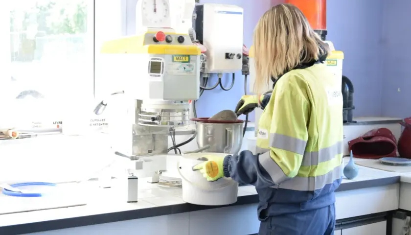 Trabajadora en laboratorio, usando guantes, manipula un recipiente con material. Se investiga qué es la silicona y sus usos en la industria.