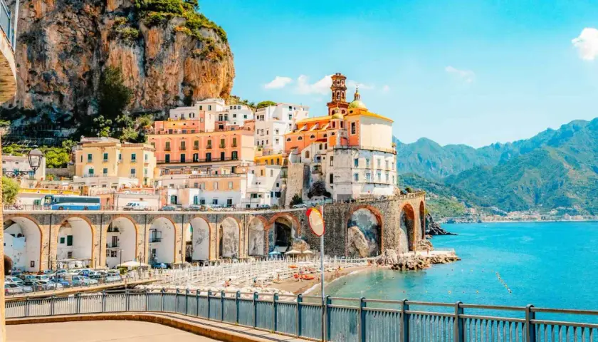 Colorful buildings cling to a cliffside overlooking a beach and the turquoise sea, a quintessential view of Amalfi Coast cities.