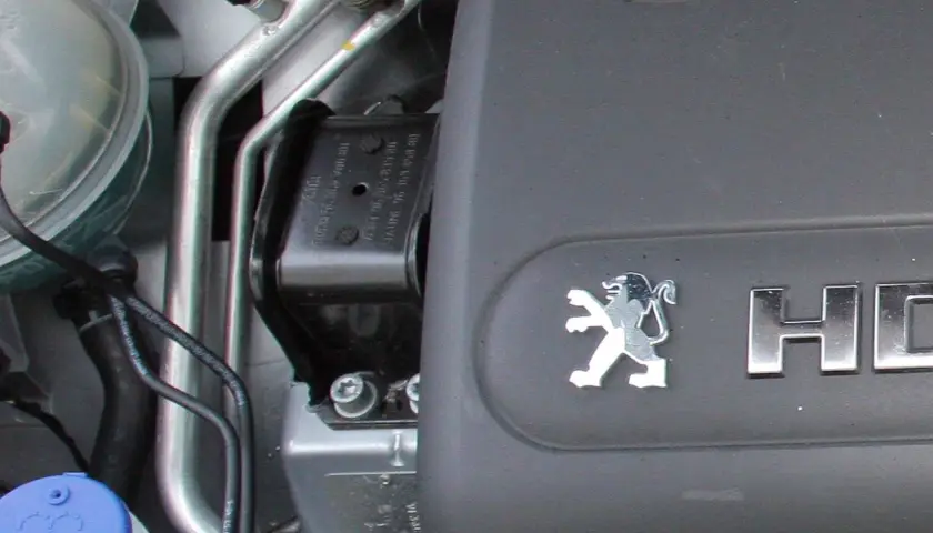 Jak sprawdzić poduszki silnika? Widok na mocowanie silnika w samochodzie Peugeot, z widocznym logo i elementami układu chłodzenia.