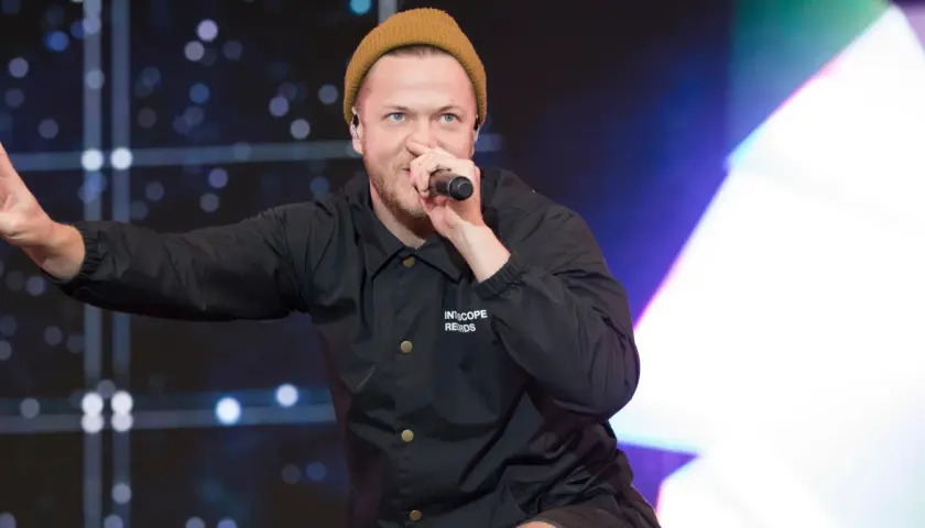 Imagine Dragons wokalista, Dan Reynolds, w beżowej czapce i czarnej kurtce, śpiewa do mikrofonu, pokazując znak pokoju.