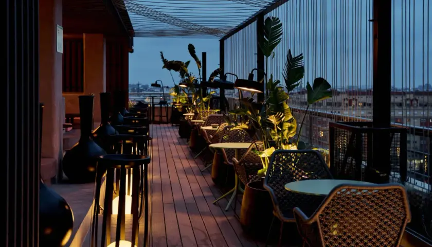 Terraza del Novotel Valencia con mesas y sillas de mimbre, iluminada por lámparas y rodeada de vegetación.
