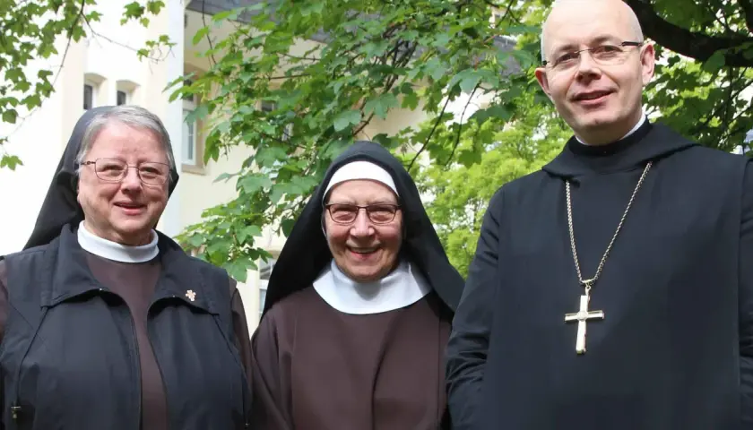 Drei Geistliche, zwei Nonnen und ein Priester, stehen lächelnd vor einem Gebäude. Sie repräsentieren katholische Orden.