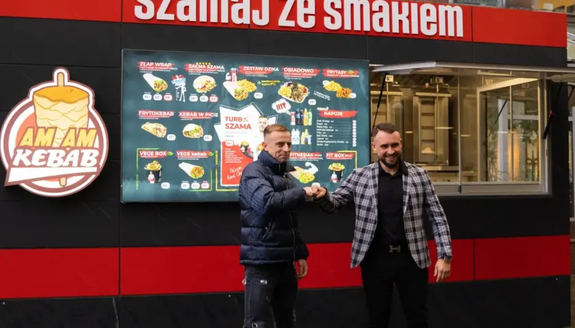 Dwóch mężczyzn wita się przy budce z kebabem, na której widnieje menu.