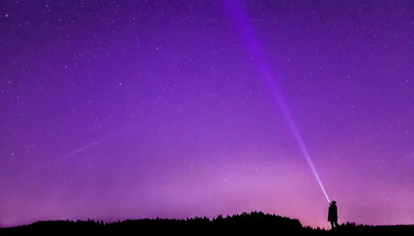 Sylwetka człowieka oświetla latarką nocne niebo, na którym widać gwiazdy i **gwiazdozbiór Tarczy**.