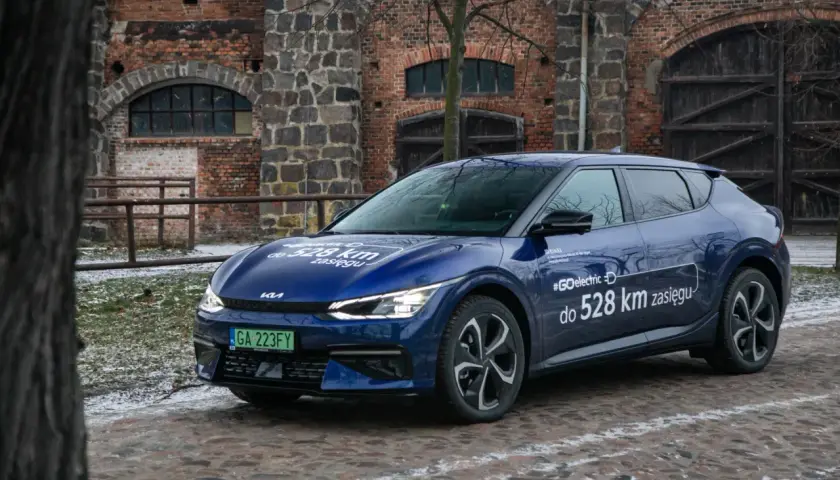 Niebieska Kia EV6 z napisem "#GOelectric D do 528 km zasięgu" stoi przed zabytkową ceglaną budowlą.
