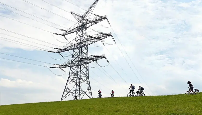 Ludzie na rowerach jadą po zielonym wzgórzu pod ogromną konstrukcją linii wysokiego napięcia.