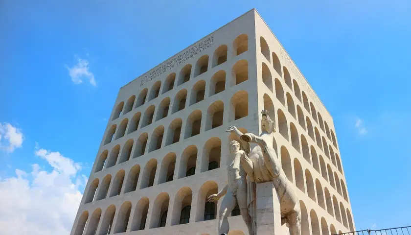 Palazzo Mussolini, con la sua facciata ad archi sovrapposti, domina il cielo azzurro. Una scultura di uomini e cavalli si erge in primo piano.