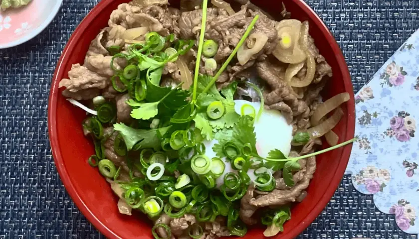 A delicious wagyu donburi with thinly sliced beef, onions, and a perfectly poached egg, garnished with fresh scallions and cilantro.
