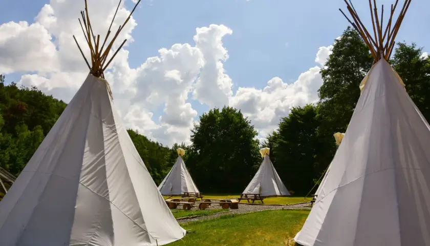 Malowniczy **pole namiotowe** z białymi tipi, drewnianymi ławami i stołami, otoczone zielenią i błękitnym niebem z chmurami.
