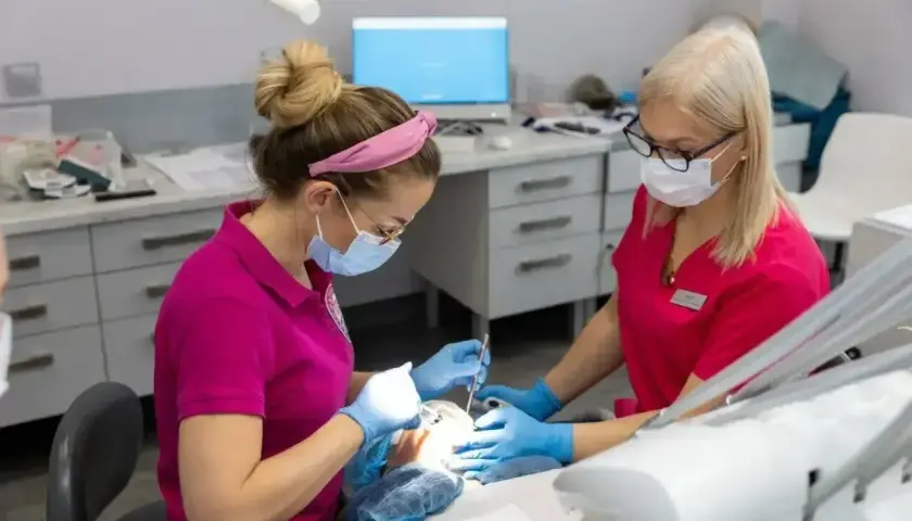 Dwie dentystki w maseczkach i rękawiczkach wykonują zabieg. Jedna z nich stosuje znieczulenie nasiękowe, by zapewnić pacjentowi komfort.