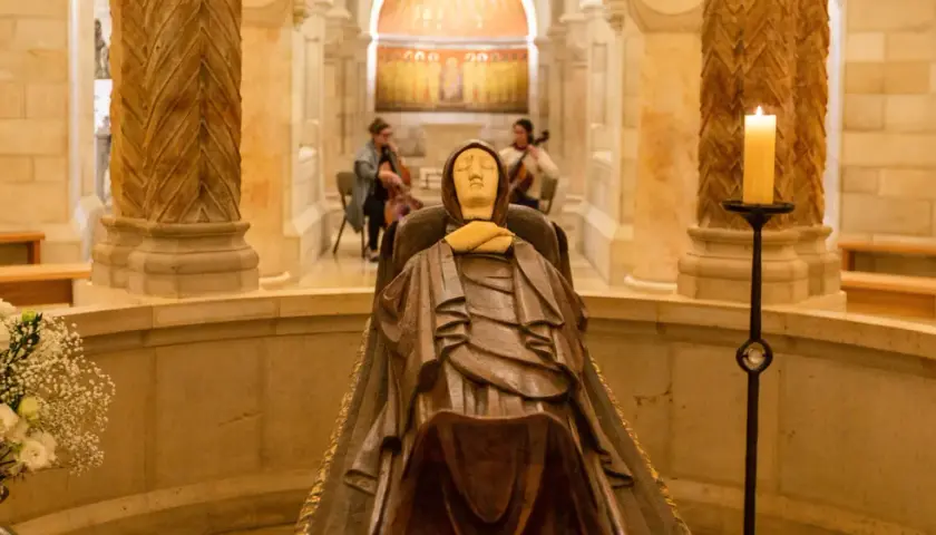 Holzschnitzerei der Mutter Gottes in einer Kapelle, mit Kerzenlicht und Blumen geschmückt. Im Hintergrund spielen zwei Musikerinnen Cello.