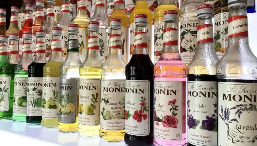Rząd butelek syropów do kawy Monin o różnych smakach: od owocowych po kwiatowe, np. róża, lawenda, hibiskus.