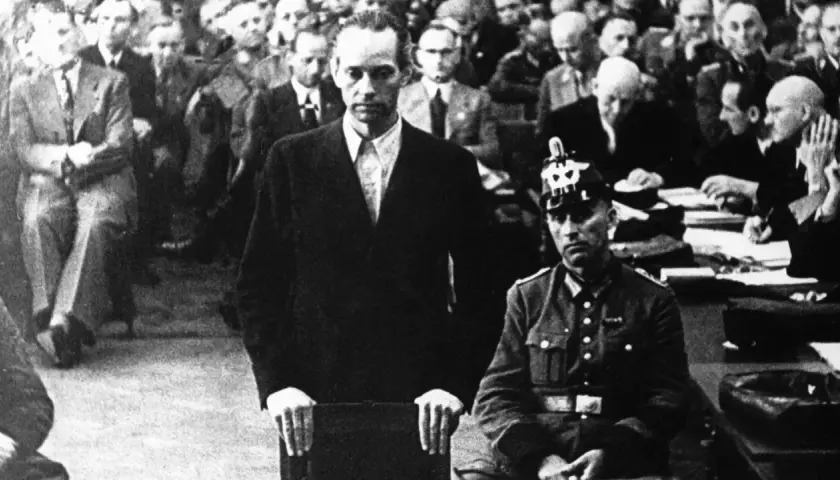 Mann im Anzug vor Gericht, umgeben von Zuschauern. Die Atmosphäre erinnert an die harten Urteile, die Roland Freisler fällte.