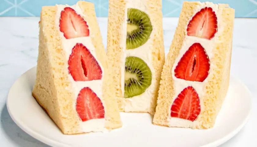 Three triangular Japanese sandwiches filled with fresh strawberries and kiwi slices, nestled in fluffy cream and soft bread.