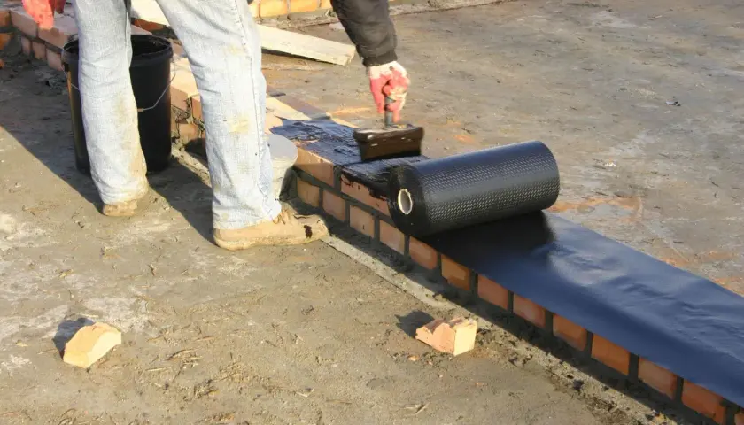 Pracownik nakłada na mur materiały hydroizolacyjne z rolki, zabezpieczając budynek przed wilgocią.