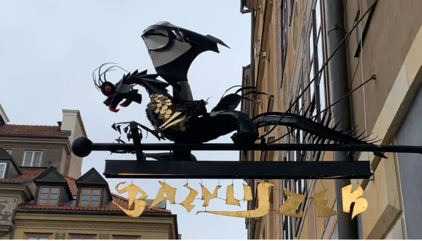 Metalowy bazyliszek ziejący ogniem wisi nad złotym napisem.