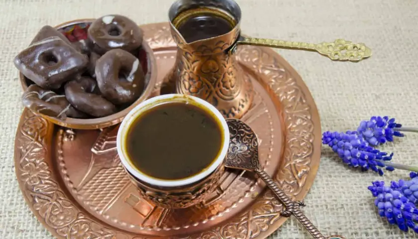 Ein traditionelles Mokkakännchen und eine Tasse mit starkem Mokka-Kaffee, dazu Schokoladenkekse und blaue Blumen auf einem Kupfertablett.