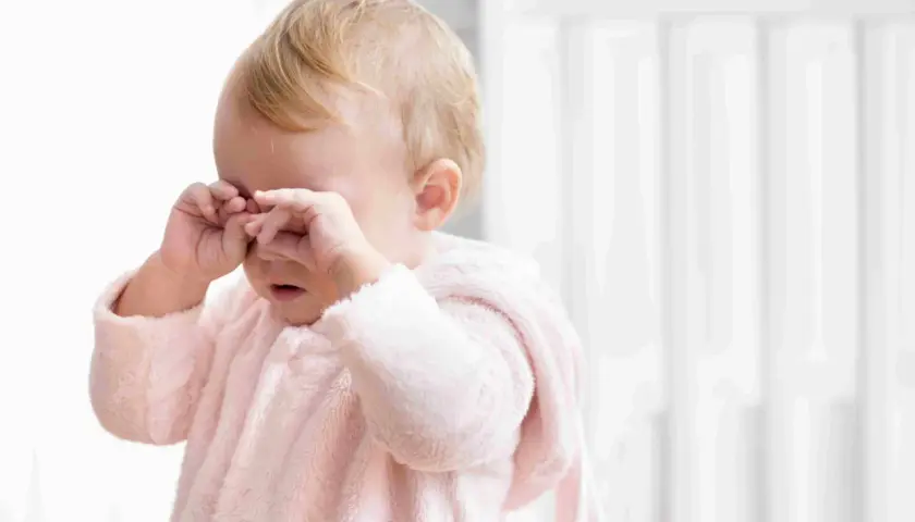 Un neonato con un pigiamino rosa strofina gli occhi, forse un occhio che lacrima per la stanchezza o un piccolo pianto.