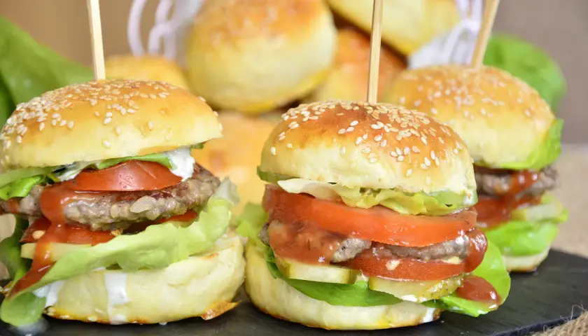 Mini burgery z sezamem, sałatą, pomidorem i sosem na czarnej desce.