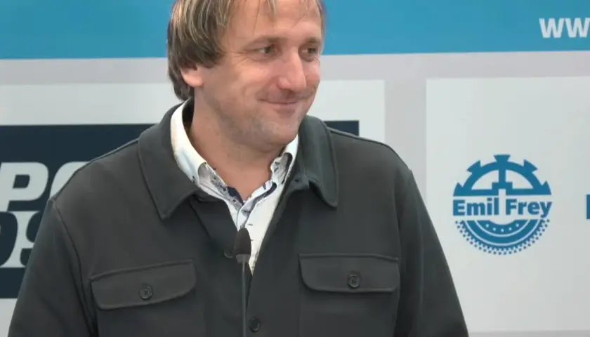 Thomas Neubert lächelt in die Kamera, trägt eine dunkle Jacke und ein gemustertes Hemd. Im Hintergrund sind Logos zu sehen.