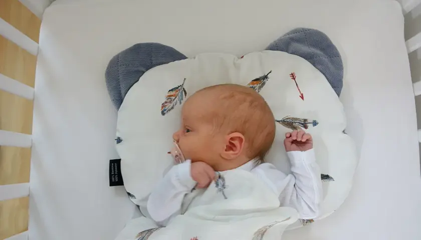 Nowy dzidziuś śpi na poduszce dla niemowlaka w kształcie misia, otulony kocykiem z piórkami.