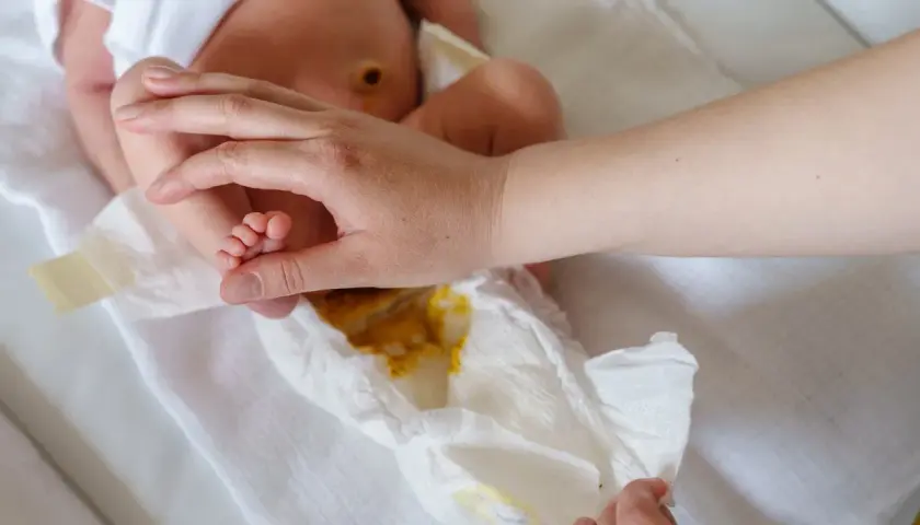 Rączka rodzica delikatnie dotyka stópki niemowlaka, podczas gdy pieluszka pokazuje zawartość – kupa niemowlaka.
