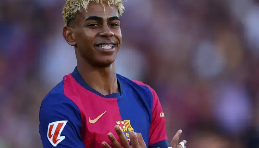Uśmiechnięty Lamine Yamal w koszulce Barcelony, z charakterystyczną fryzurą i aparatem na zębach. Jego statystyki w tym sezonie są imponujące.