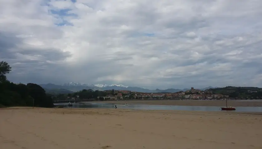 Arena dorada en la playa de la Maza, con un velero rojo en el agua y un pueblo costero al fondo, bajo un cielo nublado.