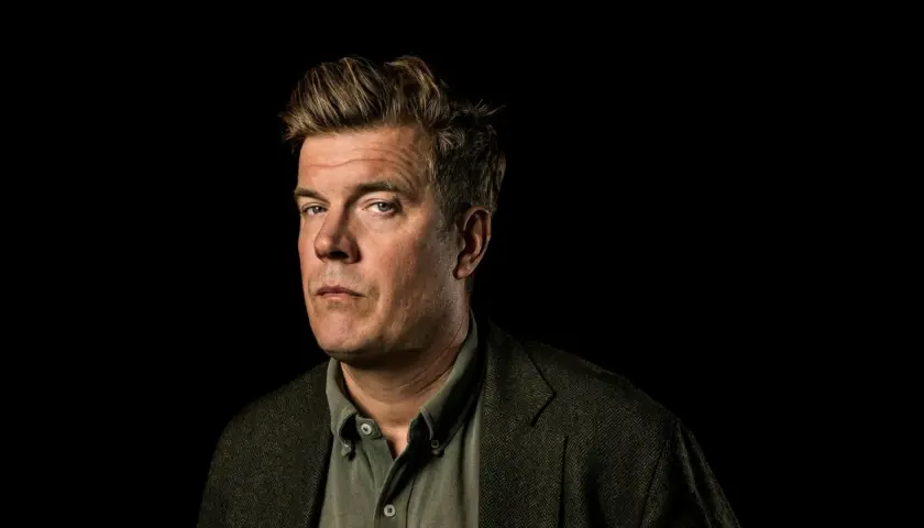 Gunnar Ardelius, en man med intensiv blick och rufsig frisyr, bär en tweedkavaj över en skjorta.