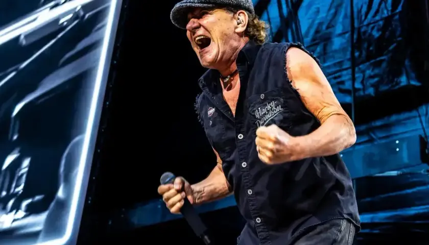 Brian Johnson z zespołu AC/DC na koncercie, w czapce i kamizelce Harley-Davidson, śpiewa z pasją do mikrofonu.